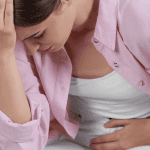 light-skinned woman with pink top on sitting on couch holding her abdomen with other hand holding head